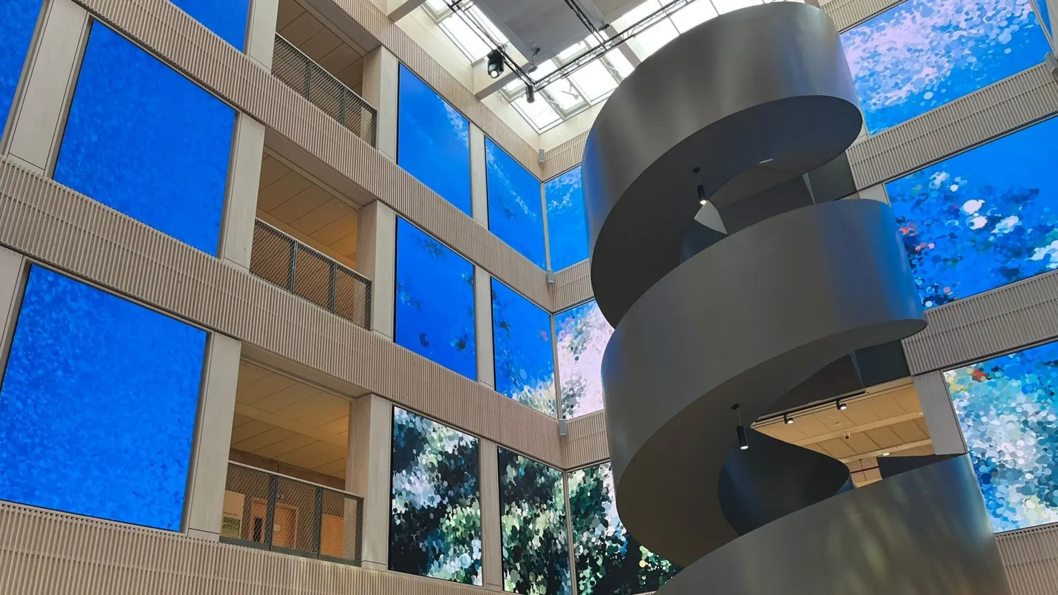 BBC / Kay Davidson A large, multi-storey indoor atrium. There is a glass roof overhead and a dark-grey spiral staircase winding up on the right. The walls surrounding are lined with tall blue rectangular panels.