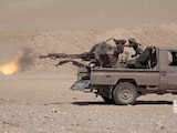 A ute with a machine gun mounted on the back tray firing in a desert.