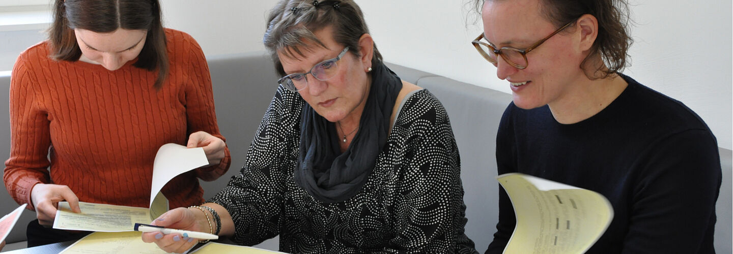 Drei Frauen an einem Tisch. Sie arbeiten mit auf dem Tisch liegenden kopierten Aktenseiten.