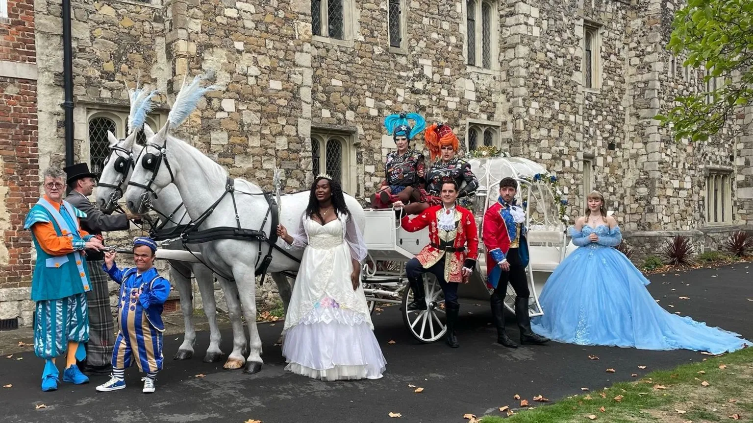 Peter Whittlesea/BBC A group of performers of a pantomime is standing outside a theatre. There is also a carriage drawn by two horses.