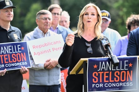 Janelle Stelson, a woman in black sweater, speaks at a podium outdoors during a campaign event, surrounded by supporters holding signs